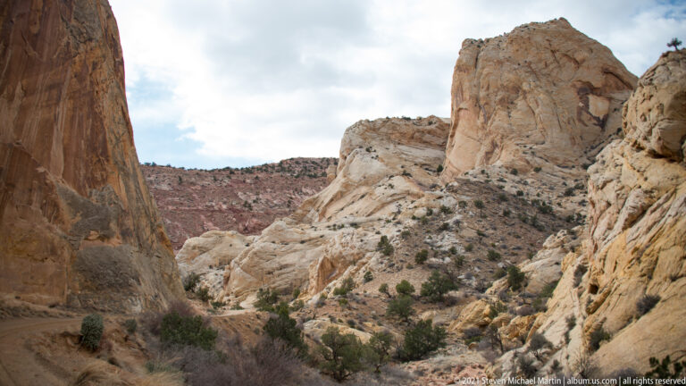 Steep pale sandstone walls frame a narrow shrub-lined drainage and a distant red ridge.