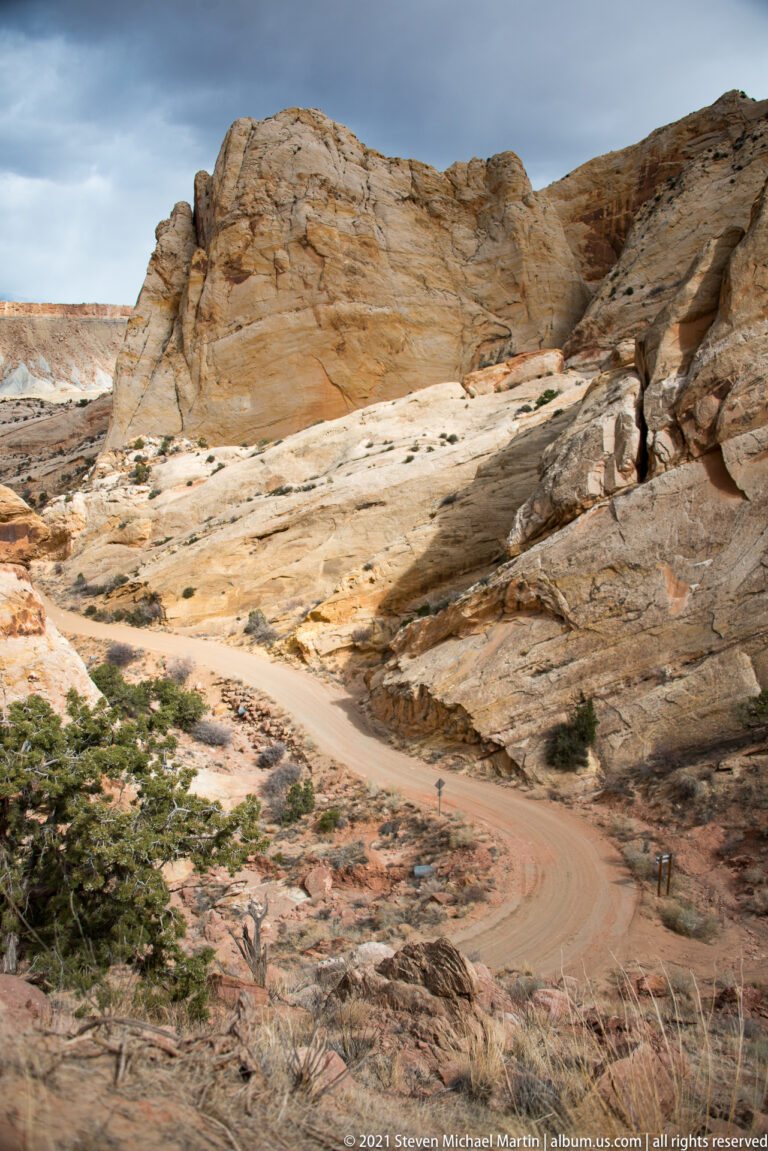 A dirt road curves through an open pale sandstone basin dotted with juniper.
