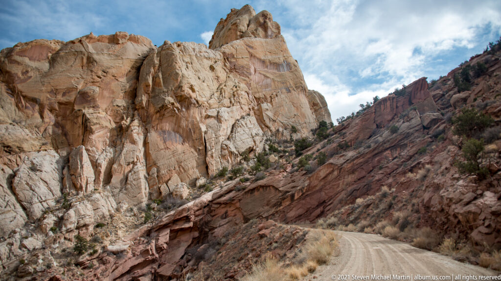 A dirt road bends below a tall sloping sandstone wall, with scrub and red outcrops nearby.