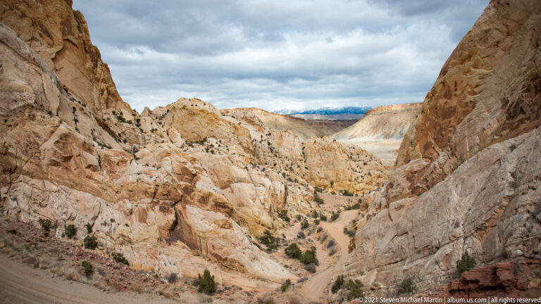 A dirt road descends between pale sandstone walls and curves toward a lower valley.
