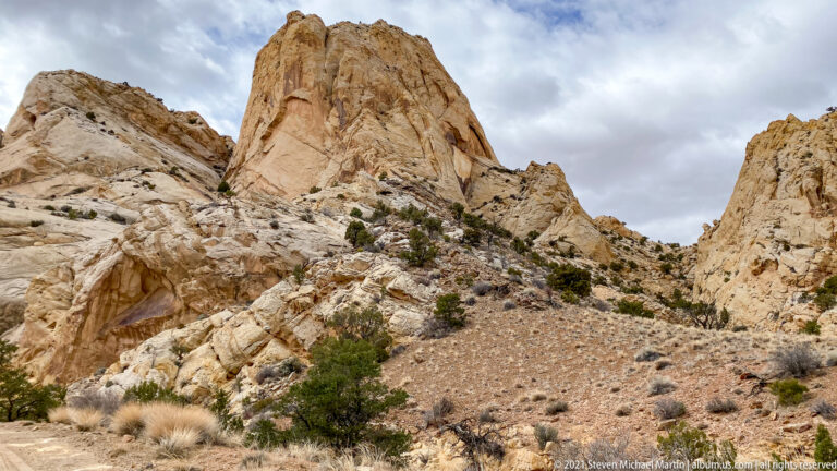 Tall cream sandstone pinnacles rise above scattered shrubs and loose rock beneath a cloudy sky.