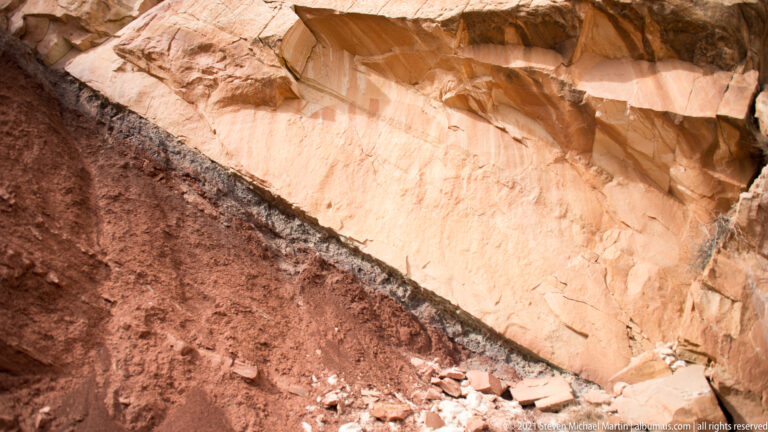 A diagonal line separates dark red rock from pale sandstone, with loose fragments below.