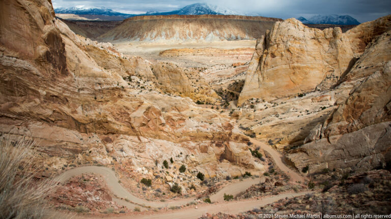 A winding dirt road crosses a pale sandstone basin beneath layered cliffs, seen from above.
