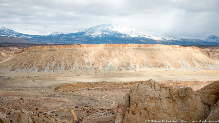 A pale gullied ridge and winding road sit below distant snow-covered mountains.