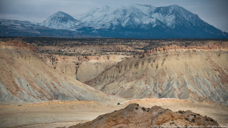 Pale eroded slopes lead toward a dark snow-covered mountain across the horizon.
