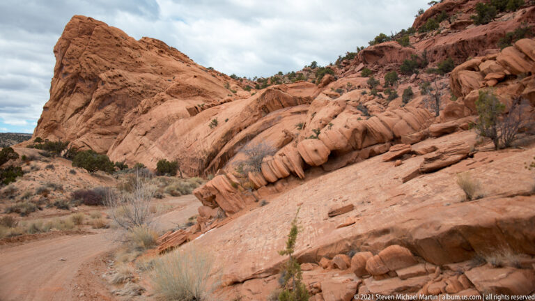 Rounded red sandstone ledges rise above a curving dirt track and dry shrubs.