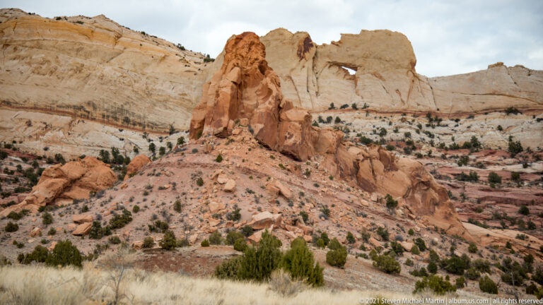 A red sandstone tower stands on a pale ridge before a layered wall with a small opening.