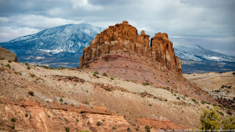 A red sandstone butte stands on a pale bench before a snow-covered mountain range.