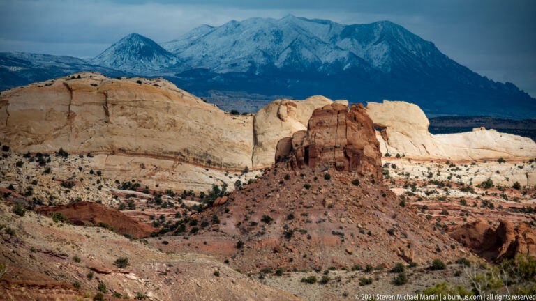 A red sandstone outcrop stands on a pale shelf below a dark snow-covered mountain.