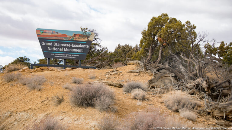 The Grand Staircase-Escalante monument sign stands among juniper and dry shrubs on pale ground.
