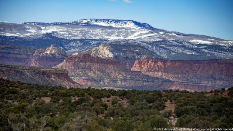 Snow covers a broad mountain above red canyon cliffs and a dark band of trees in the foreground.