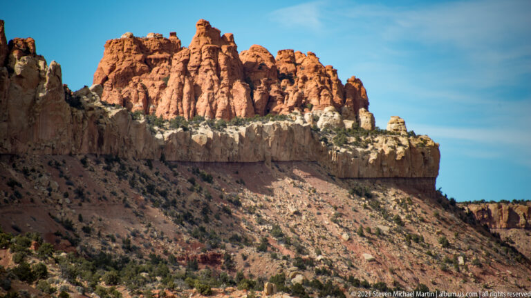 Tall red sandstone pinnacles rise above a shadowed slope beneath a clear blue sky.
