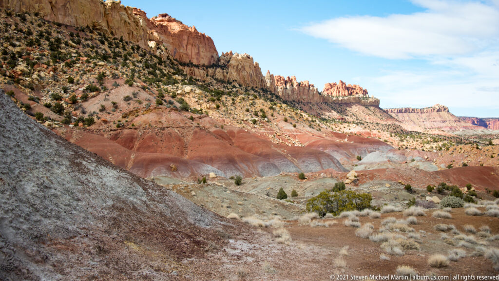 Red and pale sandstone layers sweep along a canyon slope dotted with low shrubs beneath a blue sky.