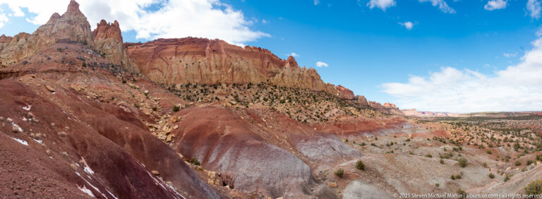 A red sandstone escarpment drops toward a distant valley, with sparse shrubs and mesas under a bright sky.