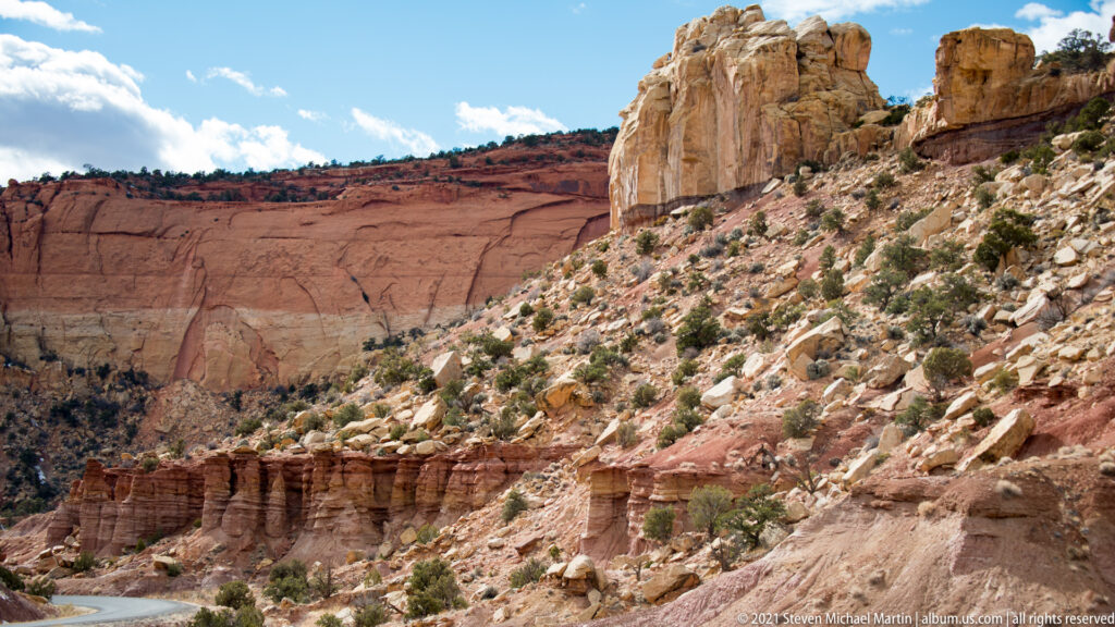 A red cliff and pale sandstone tower rise above layered shelves beside a narrow river bend.