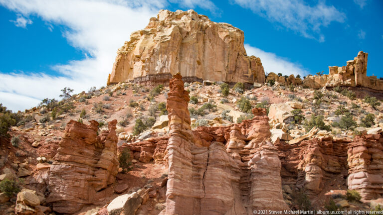 A narrow red sandstone pillar stands among fluted columns below a massive pale rock crown.