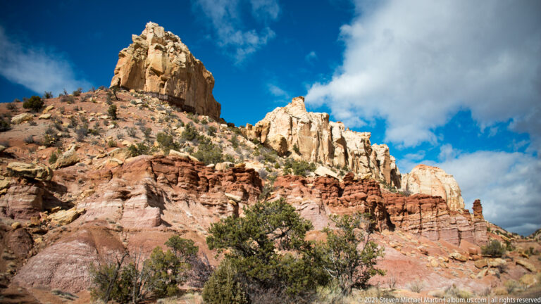 Pale towers rise above red eroded fins and shrubs beneath a bright, cloud-filled sky.