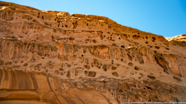 Rounded cavities and dark hollows cover an orange sandstone cliff above smoother vertical bands.