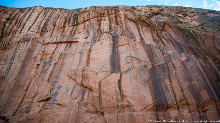 A broad sandstone face marked by parallel dark and pale bands rises toward a shrub-lined rim.