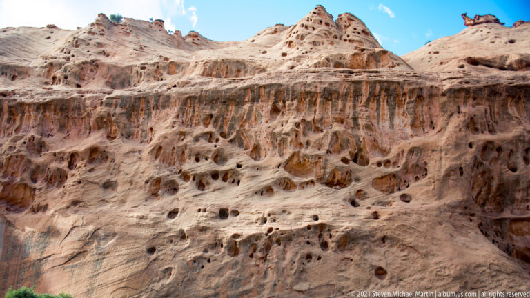 Rows of rounded cavities and dark streaks mark a pale sandstone wall beneath a sculpted ridge.
