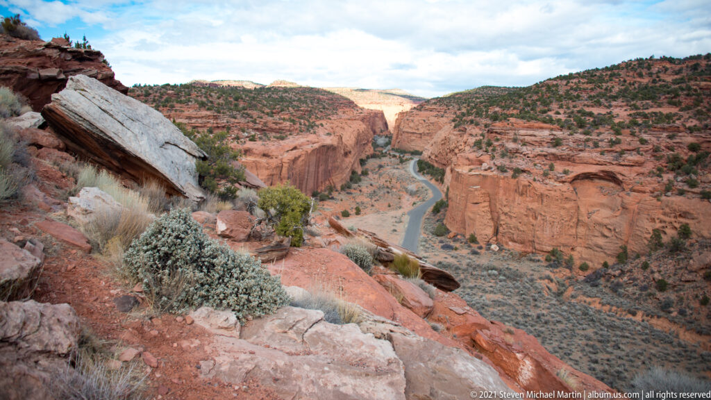 A winding canyon channel curves between steep red walls, seen from a rocky overlook with boulders in the foreground.