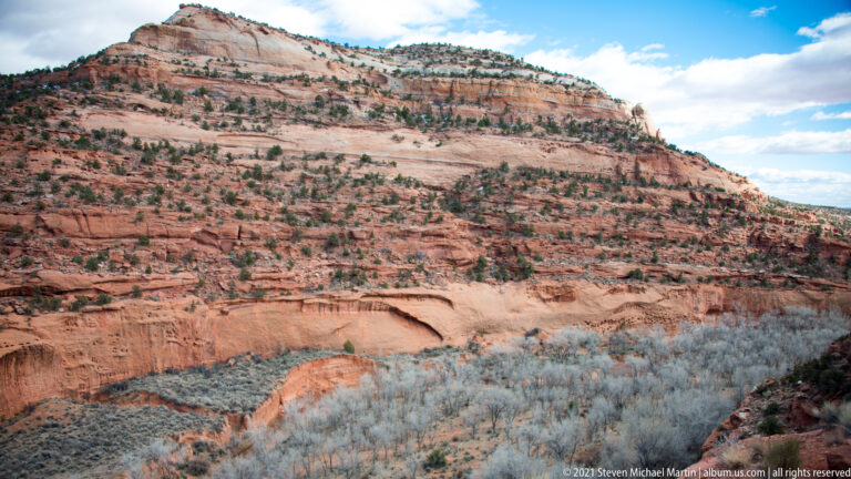 Shrub-covered slopes descend toward a deep red-rock canyon with layered benches and a winding drainage.