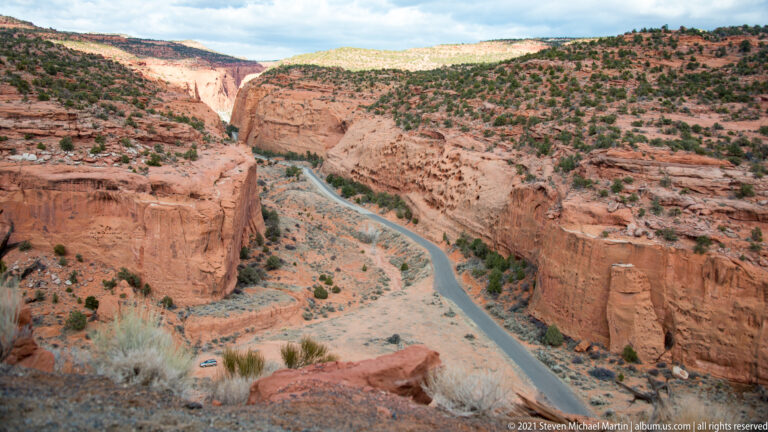 A narrow road winds along a pale canyon floor between steep red cliffs, viewed from above.