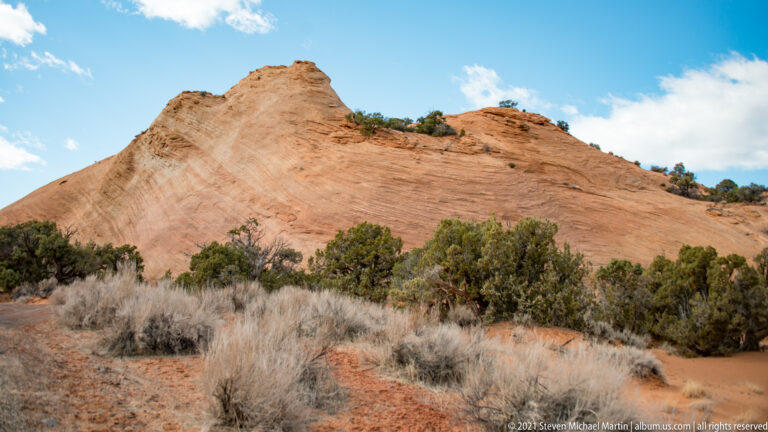 A smooth pale sandstone dome rises above red soil and sparse shrubs beneath a blue sky.