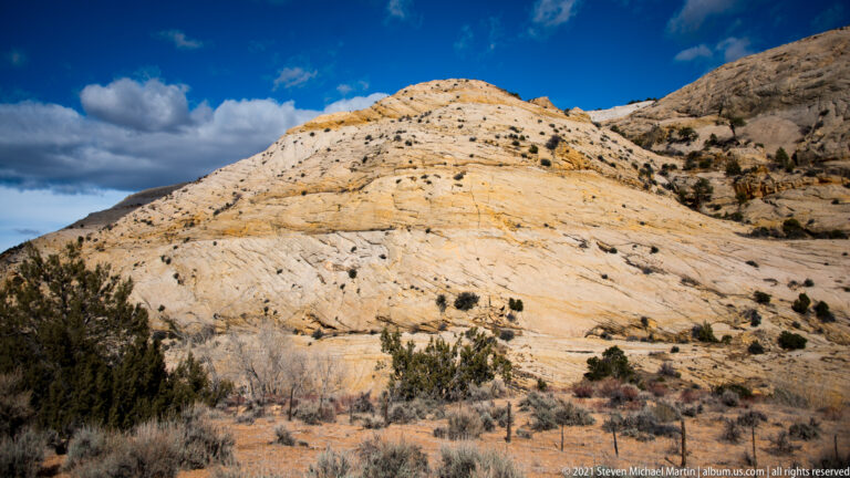 A pale sandstone slope rises above winter brush with small snow patches in its grooves and ledges.