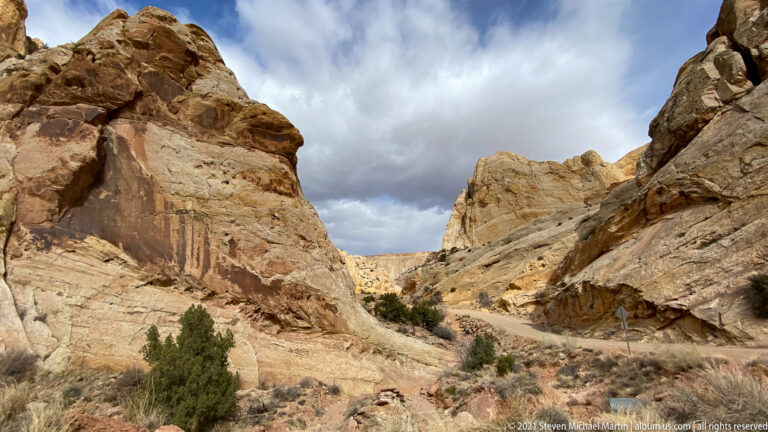 A narrow dirt road curves between high sandstone walls, with a small sign at the roadside.