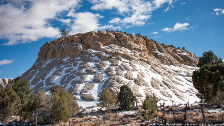 White snow traces grooves across a rounded sandstone dome framed by shrubs beneath a blue sky.