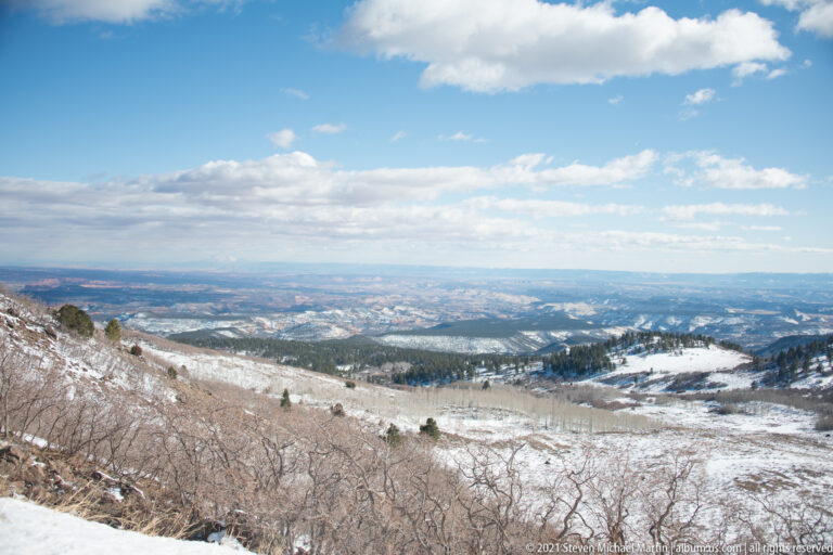 Snow and bare shrubs cover a ridge above a broad valley with distant roads and settlements.