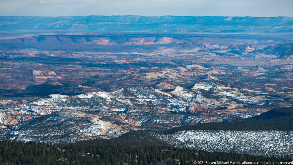 Snowy ridges and dark forest overlook red canyon walls and a distant plateau.