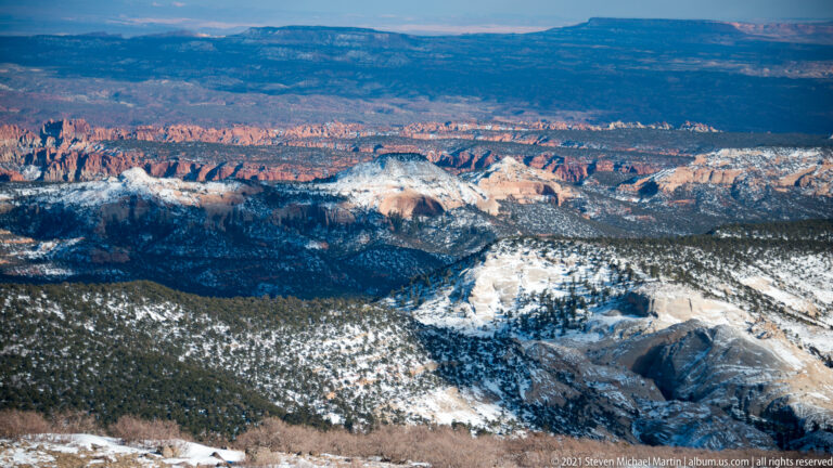 Snowy high ground overlooks forest, red canyon cliffs, and distant plateaus under patchy sunlight.