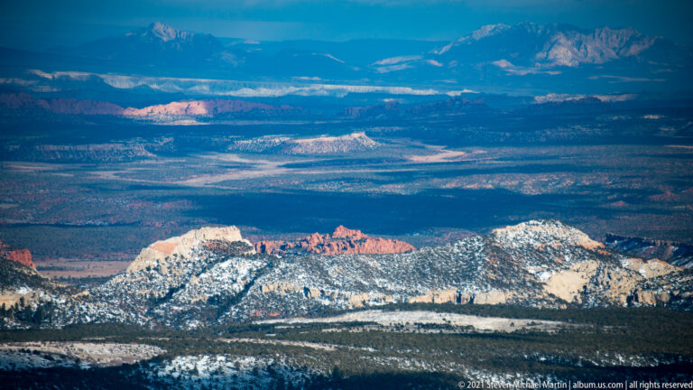 Snowy forest and red sandstone ridges form layers below distant blue-gray plateaus and mountains.