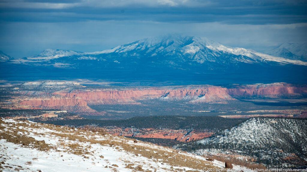 A snow-covered mountain rises above a red canyon rim, with snow on the nearer slope and cloud around the peak.