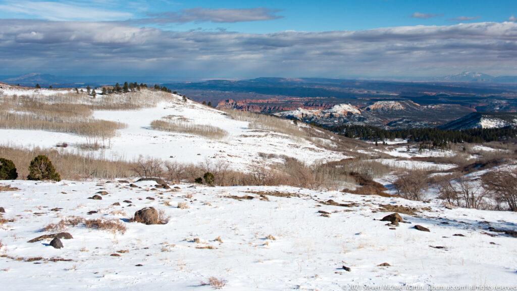 A snowy slope with scattered rocks overlooks forest and distant red canyon walls beneath a long cloud band.