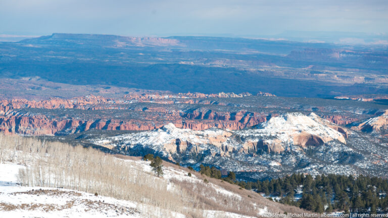 Snowy ridges and forest lead toward red canyon cliffs and pale plateaus in a broad basin.