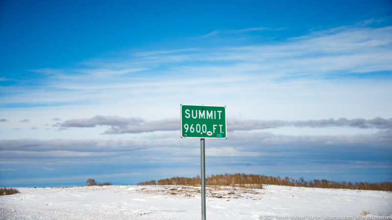 A green sign reading “Summit 9600 ft” stands in a snowy clearing beneath blue sky and a distant cloud band.