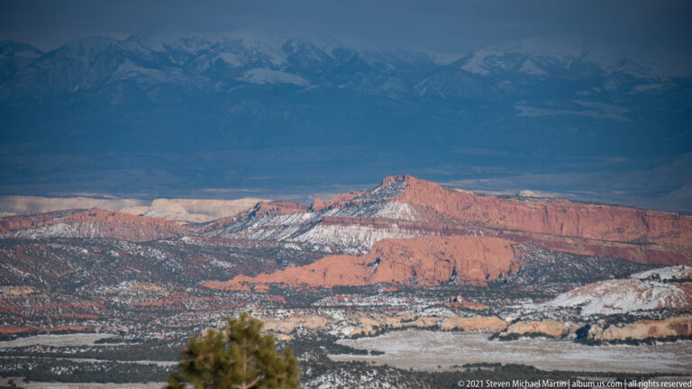 Snow traces pale lines across red-rock ridges, with a hazy mountain range behind them and a small tree at the bottom edge.