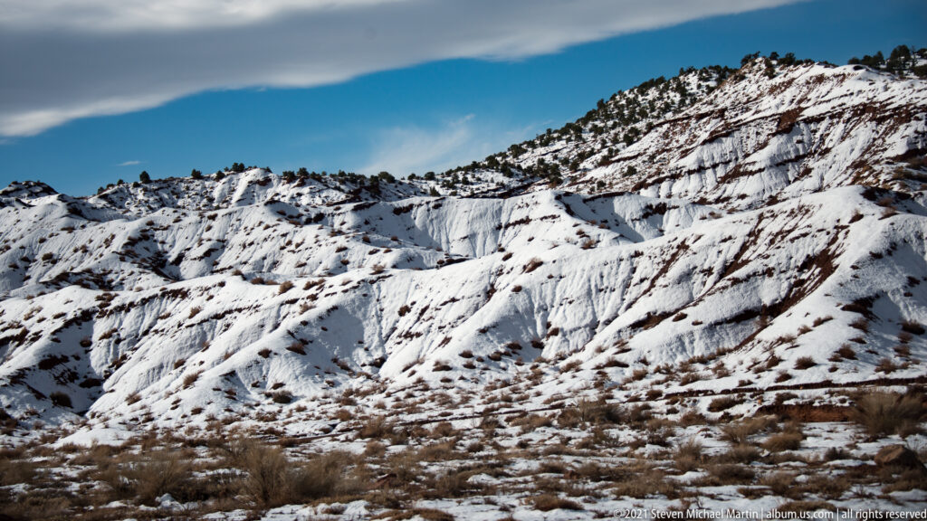 Snow traces ledges and gullies across layered sandstone slopes, with sparse shrubs below a blue sky.