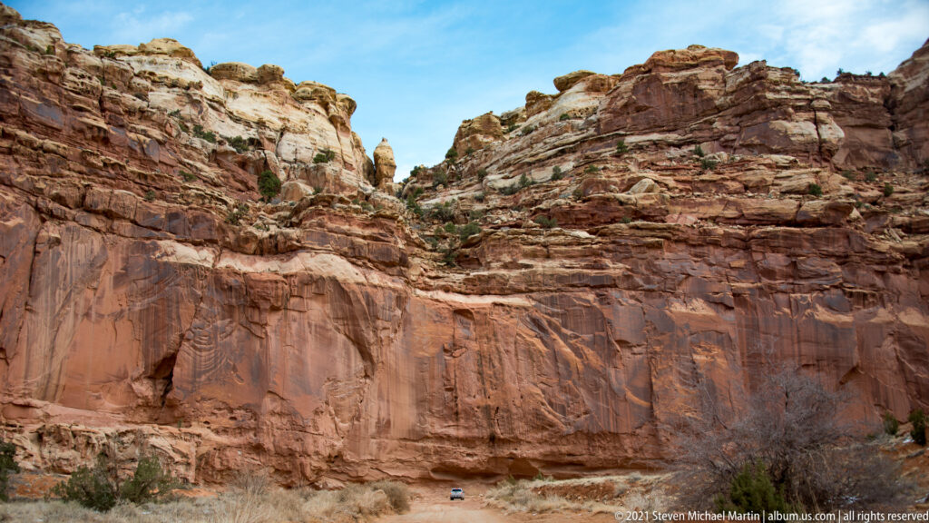A small vehicle sits in a rocky wash between tall, layered red and tan canyon walls.