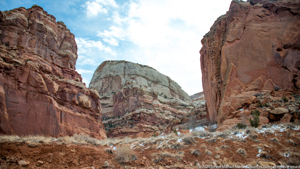A pale rounded rock dome appears between red canyon walls above a snow-dusted foreground.