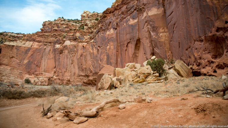 A tall red sandstone wall stands above a dry wash and pale fallen boulders.