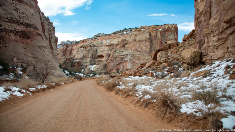 A dirt road leads between red and cream canyon walls, with snow patches along the brushy edges.