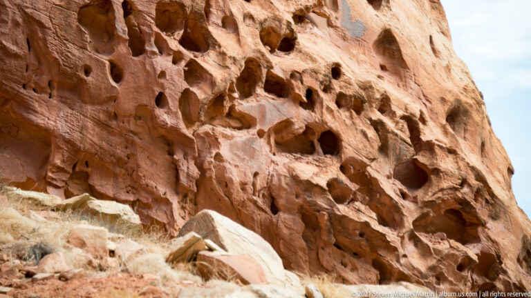 Rounded holes and shadowed alcoves cover a red sandstone wall above scattered rock and dry grass.