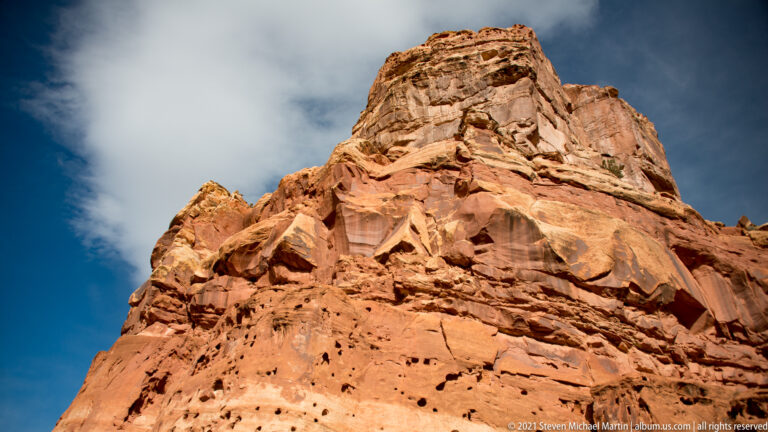 Low-angle view of a tall layered sandstone buttress with ledges and small shrubs against blue sky.