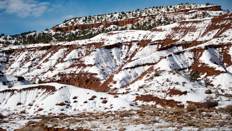 Broad snow-covered ridges show horizontal red bands and scattered shrubs beneath a clear sky.