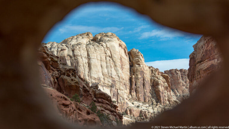 A rounded dark rock opening frames pale sandstone cliffs and blue sky in the distance.