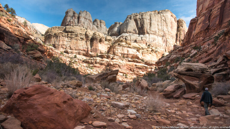 A lone hiker crosses a rocky wash toward pale cliffs between red canyon walls.